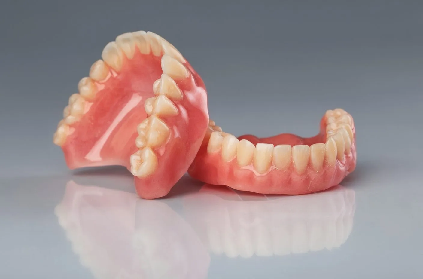 Upper and lower full acrylic dentures on grey reflective surface; studio product shot; warm pink acrylic base