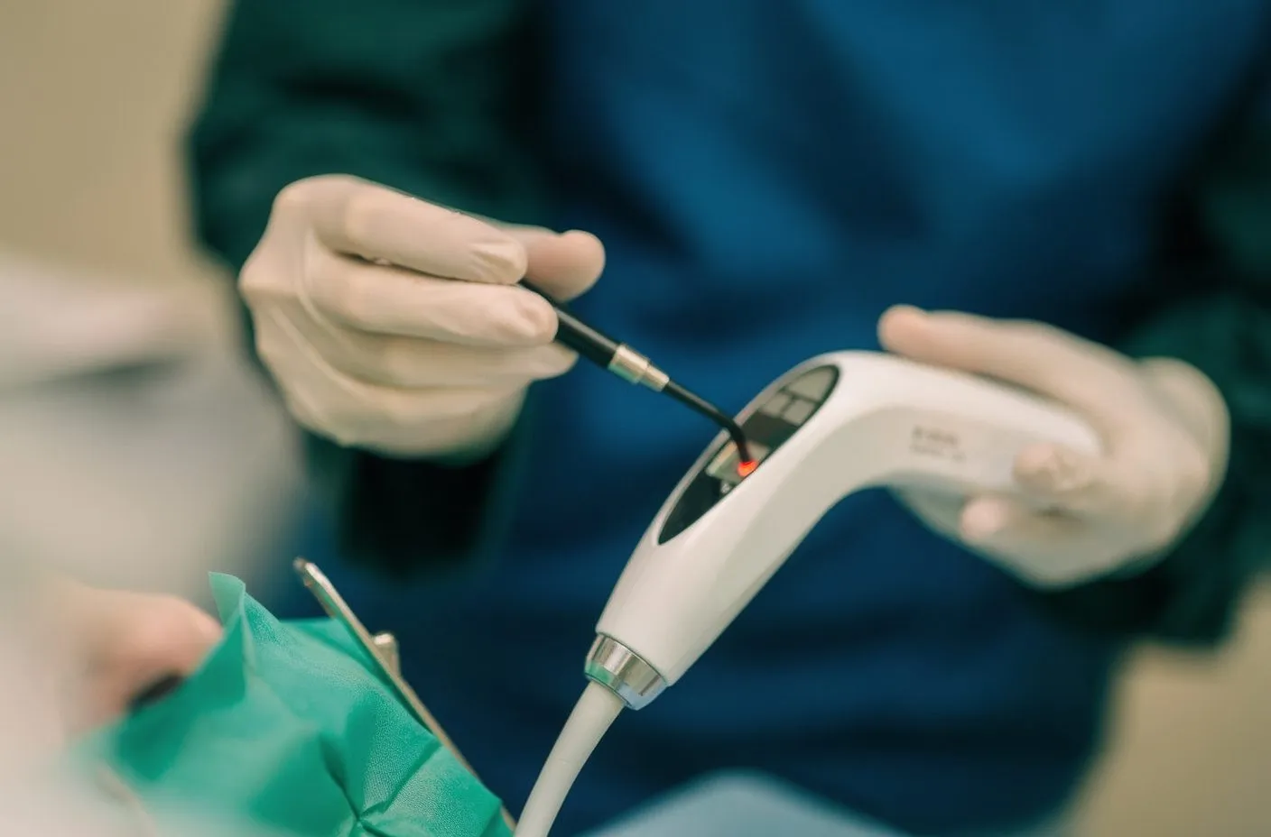 Gloved hands holding dental laser handpiece with red indicator light and fiber-optic tip; green surgical drape
