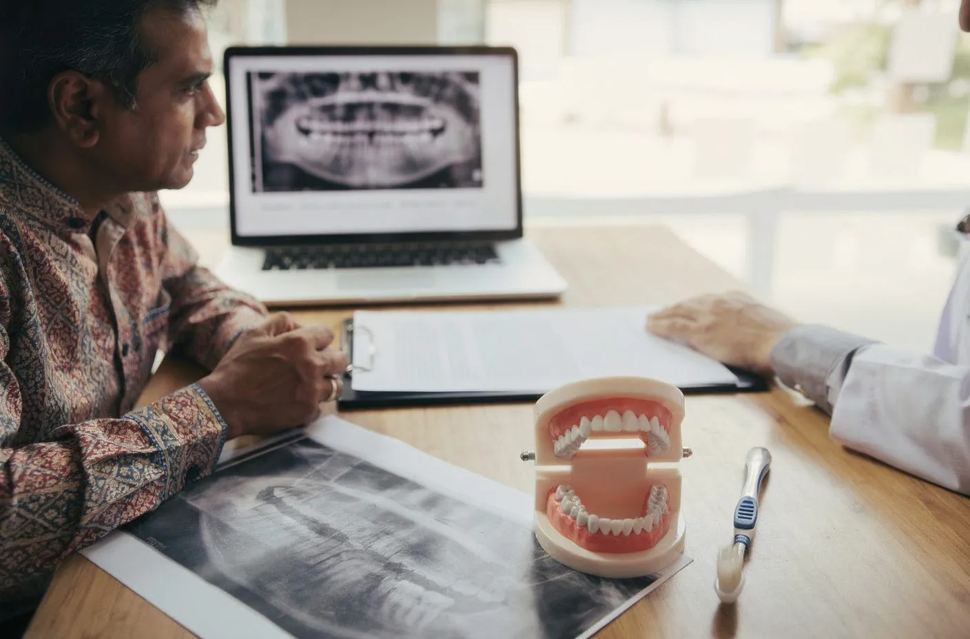 South Asian male patient in consultation with dentist; jaw model and X-ray on desk, panoramic on laptop