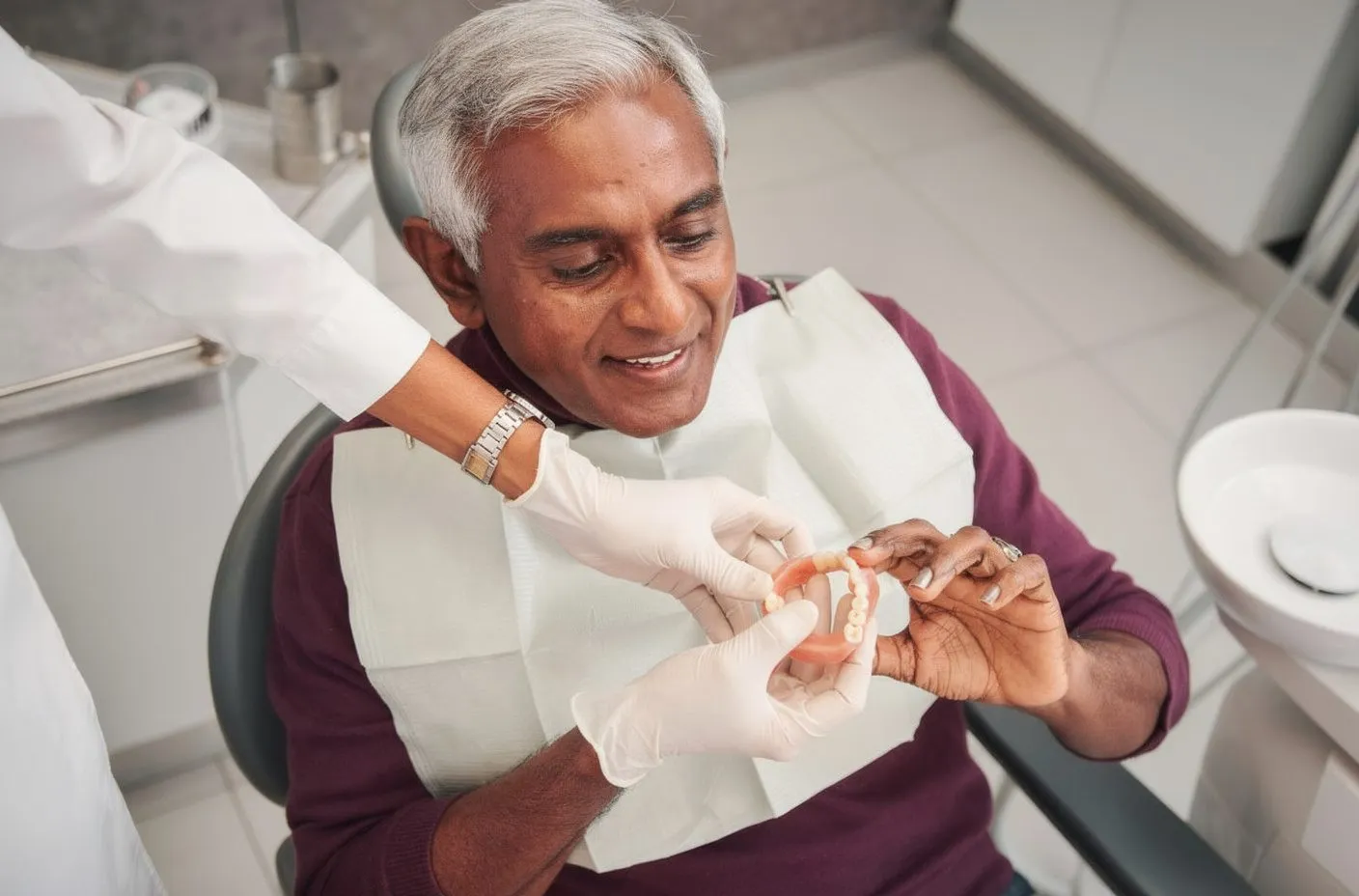 Middle-aged South Asian man in dental chair; dentist hands him a denture to examine; warm, reassuring mood